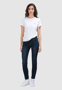 Wit katoenen t-shirt met een rood grafisch ontwerp op de linkerborst, gecombineerd met donkerblauwe skinny jeans en witte hoge sneakers.