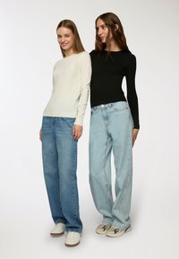 Dwie modelki noszą dopasowane bluzki z długim rękawem: jedna w kolorze kremowym, a druga w czarnym. Dodatkowo, każda z nich ma na sobie szerokie dżinsy, odpowiednio jasnoniebieskie i ciemnoniebieskie.