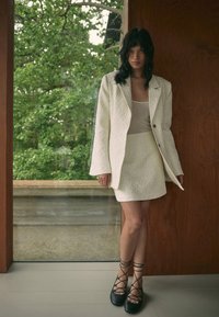 Blazer blanc texturé et jupe assortie, accompagnés de chaussures noires à brides. L'environnement est baigné de lumière naturelle avec de la verdure visible à travers la fenêtre.