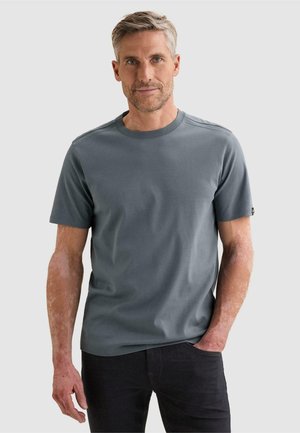 Homme aux cheveux courts gris portant un t-shirt gris uni à manches courtes et un pantalon noir, debout avec une main dans la poche sur un fond neutre.