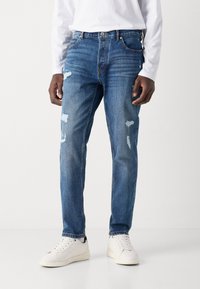 Siniset farkut slim-fit tyyliin, joissa on kulutuspisteitä ja repaleisia yksityiskohtia, yhdistettynä valkoisiin tennareihin ja pitkähihaiseen valkoiseen paitaan.