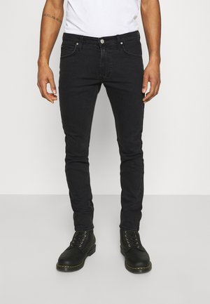 Man wearing black slim-fit jeans, black lace-up boots, and a white t-shirt, standing against a plain light background.