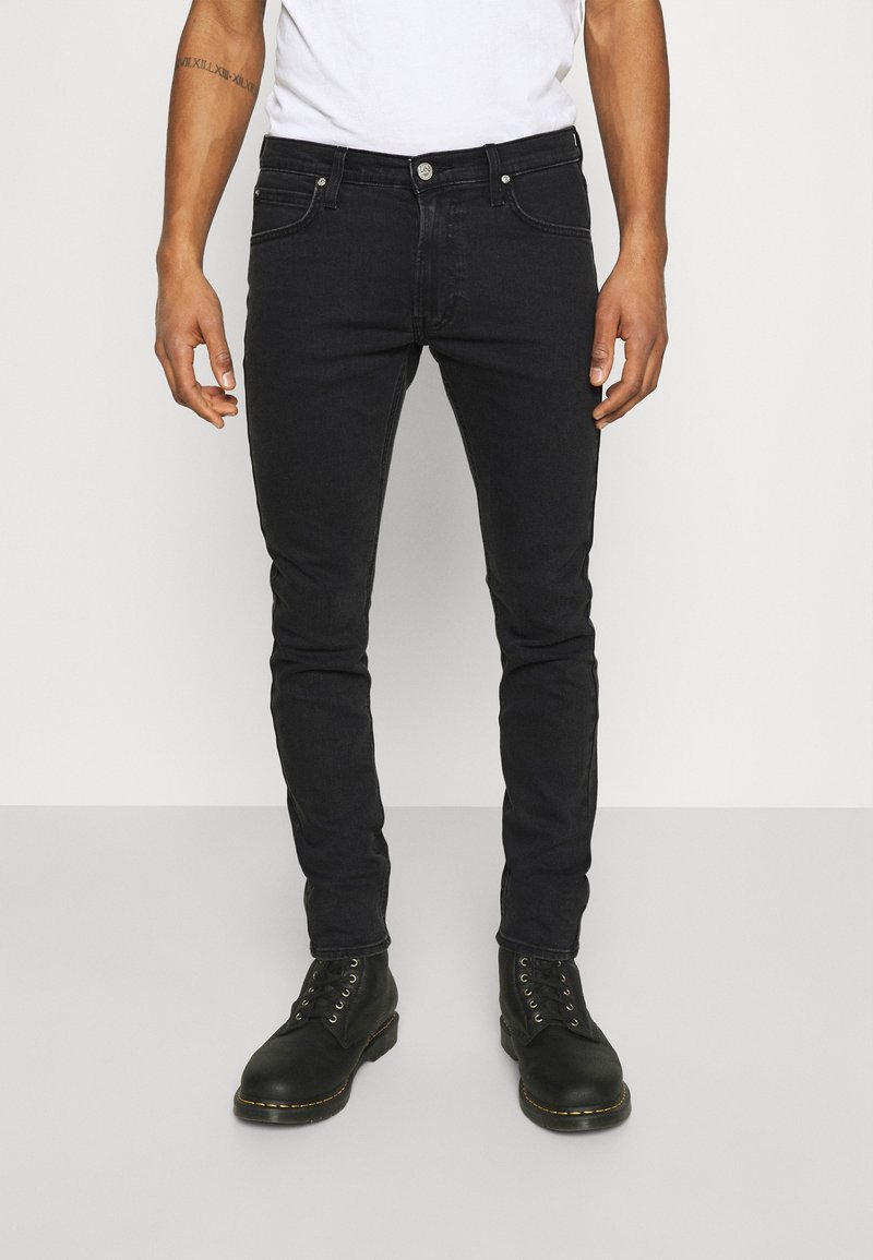 Homme portant un jean noir ajusté, des bottes noires à lacets et un t-shirt blanc, debout contre un fond clair uni.