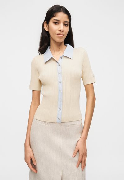 Γυναίκα που φοράει ένα μπεζ κοντομάνικο ριμπ δετό ζακέτα με γαλάζιο ριγωτό γιακά και κουμπότρυπες, σε συνδυασμό με μια μπεζ φούστα.