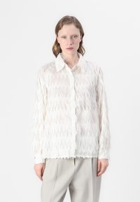 Blusa bianca con frange, caratterizzata da un motivo testurizzato, dotata di colletto classico e chiusura con bottoni. Abbinata a pantaloni grigi chiari.