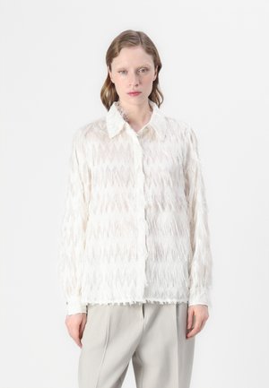 Blusa bianca con frange, caratterizzata da un motivo testurizzato, dotata di colletto classico e chiusura con bottoni. Abbinata a pantaloni grigi chiari.