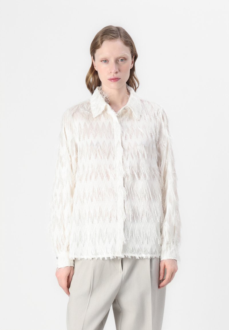 Blusa bianca con frange, caratterizzata da un motivo testurizzato, dotata di colletto classico e chiusura con bottoni. Abbinata a pantaloni grigi chiari.