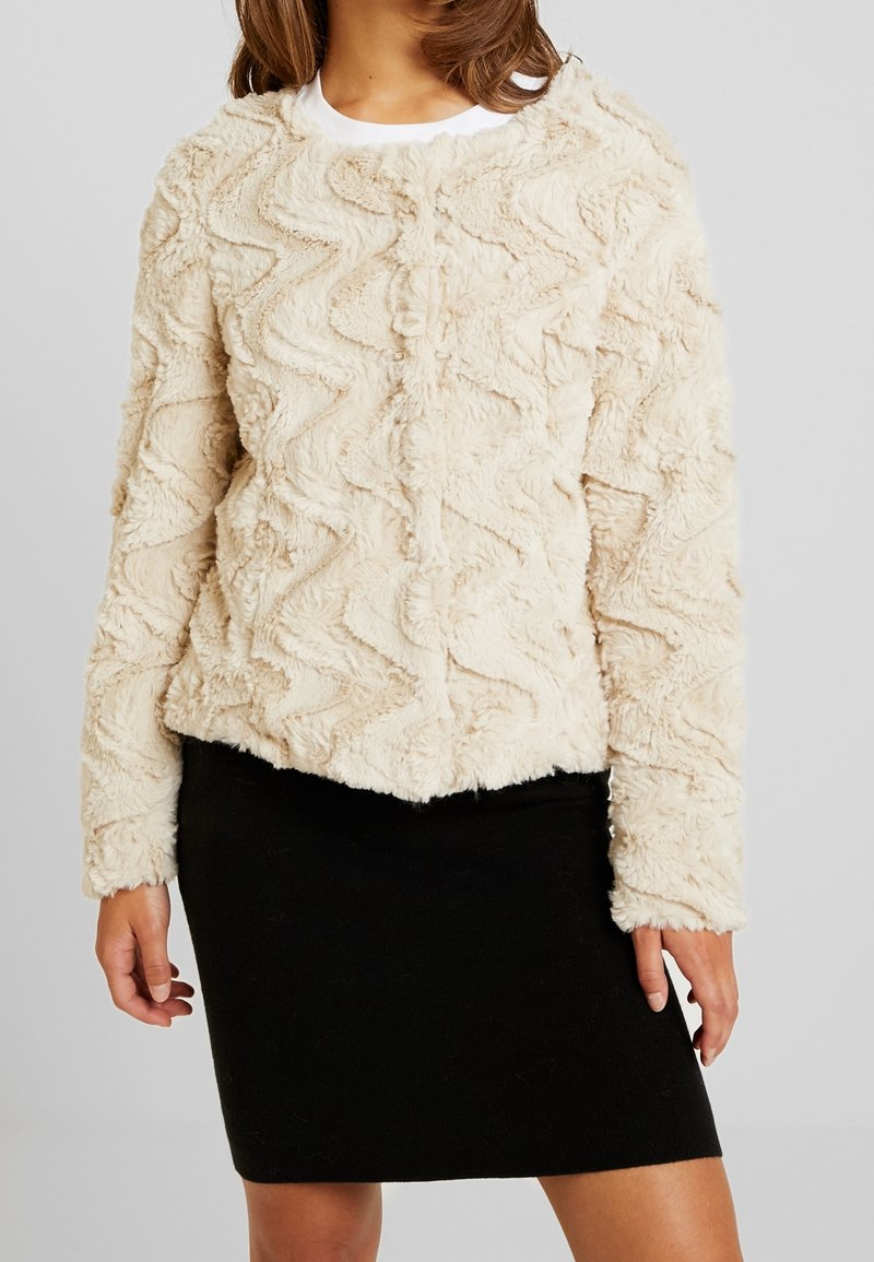 Beige väriltään teksturoitu tekoturkistakki, jossa on siksak-kuviot, pyöreä pääntie ja pitkät hihat, yhdistetty mustaan kynähameeseen.