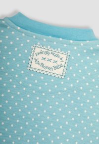 Hellblauer Baumwollstoff mit weißen Punktmotiven. Enthält ein gesticktes Etikett mit Text in kontrastierender Farbe. Weiche, glatte Textur.