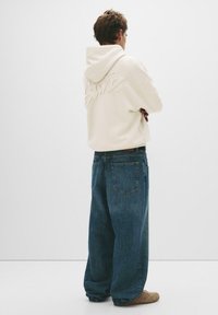 Cremefarbener Hoodie mit gesticktem Text auf dem Rücken, kombiniert mit lockeren, dunkelblauen Jeans und beiger Fußbekleidung. Minimalistisches Design.