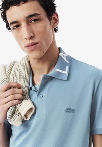 Jasnoniebieska koszulka polo o teksturowym wykończeniu, z białym logo na kołnierzyku oraz małym haftowanym logo krokodyla. Biały sweter dzianinowy zarzucony na ramię.