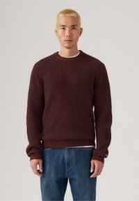 Burgundy knit sweater with a textured surface, crew neckline, and ribbed cuffs. Paired with blue denim jeans, featuring a relaxed fit.