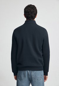 Sudadera azul marino con cuello y puños acanalados; tela suave; la vista trasera muestra hombros estructurados y un ajuste relajado, combinada con unos pantalones vaqueros de mezclilla claros.