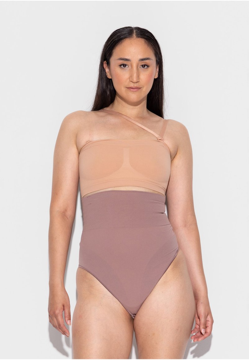 Moteris su ilgais tamsiais plaukais vilki dvispalvį maudymosi kostiumėlį su viena petneša, stovinti priešais šviesiai pilką vientisą foną, žiūrinti tiesiai į priekį.