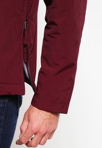 Bordeaux jas van een gladde, glanzende stof; voorzien van een rits en een grijze binnenvoering. De uiteinden van de mouwen hebben een nette afwerking met stikseldetail.