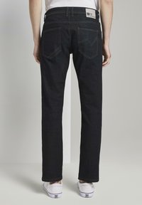 Vaqueros de mezclilla oscura de corte recto, con bolsillos traseros distintivos y una etiqueta de marca en la cintura. Zapatillas visibles en los tobillos.