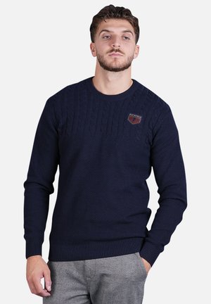 Pull en tricot bleu marine avec un motif de câble texturé, des poignets et un ourlet côtelés, arborant un petit écusson logo sur la poitrine.