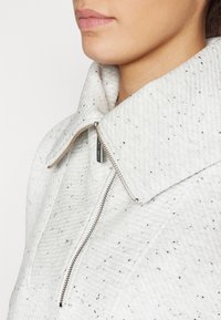 Λευκή επενδυμένη ζακέτα με μεγάλο γιακά, με μαύρες πιτσιλιές, μεταλλικό φερμουάρ και υφή στην επιφάνεια.