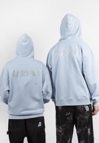 Vaaleansiniset hupparit, joissa suuri strassitekstigrafiikka takana. Pehmeä kangas, etupuolella kengurutaskut, yhdistetty mustiin kuluneisiin housuihin.