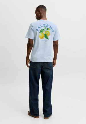 Mand iført en lyseblå Palermo t-shirt med citronmotiv og mørkeblå jeans, stående med ryggen mod en ensfarvet baggrund.