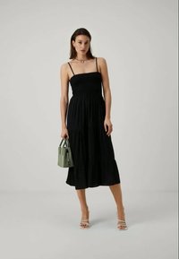 Čierne šaty na tenkých ramienkach s vrstveným, splývavým dizajnom. Modelka drží malú zelenú kabelku a má na sebe biele sandále na remienky. Jednoduché pozadie.