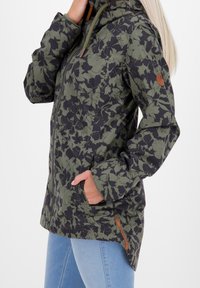 Vrouw in een groen en zwart bloemenpatronen jacket met rits en zijzakken, gecombineerd met lichtblauwe jeans, met haar handen in de zakken.
