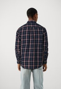 Camicia a quadri blu navy con motivi rossi e bianchi, polsini con bottoni e un dettaglio di yoke sul retro. Tessuto morbido, maniche lunghe, orlo leggermente curvo.