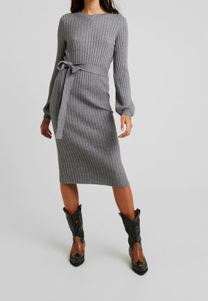 Woman wearing a grey ribbed knit midi dress with a tied waist belt and black and brown cowboy boots, standing on a grey floor.