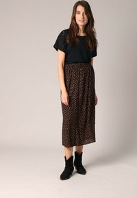 Femme se tenant debout, portant un haut noir à manches courtes, une jupe midi noire à pois orange et des bottes noires à chevilles, contre un fond uni.