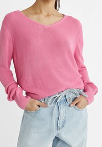 Pull en maille rose à col en V avec texture côtelée, manches longues et coupe décontractée, porté avec un jean en denim bleu clair.