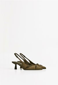 Tacchi slingback in suede verde oliva con punta affilata, cinturini sottili e un piccolo tacco a blocco. Dettaglio notevole: accentuata fibbia decorativa.