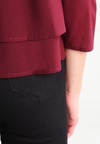 Top bourgogne en tissu léger avec un design superposé. Jean noir avec une poche arrière en forme de losange. Texture lisse, silhouette ajustée.