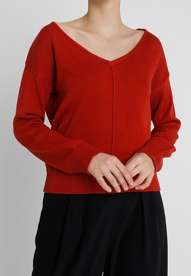 Femme portant un pull en maille rouge à col en V, manches longues avec poignets et ourlet côtelés, associé à un pantalon noir ample.