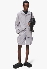 Jasnoszara, wodoodporna kurtka z kapturem, zapięciem na guziki i kieszeniami z przodu; dopasowane szorty z kieszeniami cargo; masywne czarne obuwie.