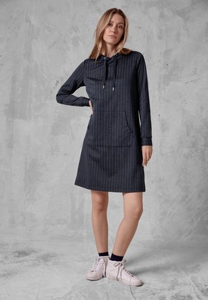 Femme debout, main sur la hanche, portant une robe à capuche rayée foncée et des baskets montantes blanches, devant un fond texturé gris.