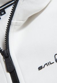 Chaqueta blanca con cremallera y logo "SAIL" en negro. La tela es suave, con un cuello y detalle de franja azul en el interior.
