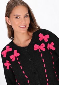 Juodas megztinis su rožinėmis juostelėmis ir kontrastinėmis siuvimo detalėmis, su juodais mygtukais priekyje.