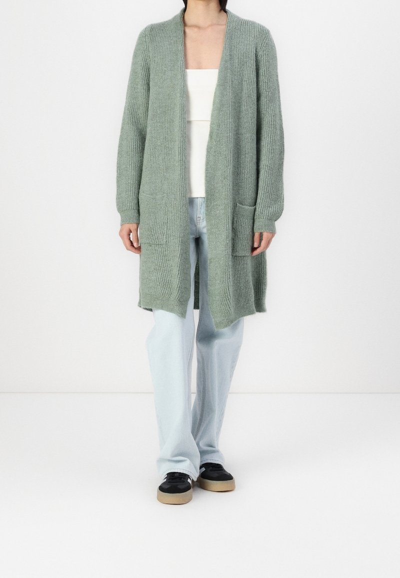 Personne portant un long cardigan gris en maille, un haut blanc, un jean bleu clair et des baskets noires avec des semelles beige, debout devant un fond blanc.