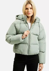 Kobieta z długimi, blond włosami, ubrana w jasną zieloną kurtkę puchową z kapturem, zapięta na zamek nad białą bluzką i czarnymi spodniami.