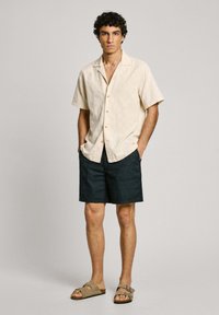 Camisa corta de manga corta en beige claro con una sutil textura, combinada con pantalones cortos azul marino oscuro. Sandalias marrones completan el look. Diseño simple y casual.