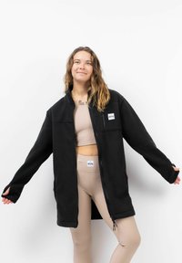 Zwarte fleecjacket met een ritssluiting, twee frontzakken en ribbelafwerking bij de hals en mouwen. Een lichte achtergrond accentueert het ontwerp.