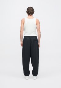 Juodos laisvo silueto sportinės kelnės su elastine liemene ir užpakaline kišene. Derinamos su baltu rankovių marškinėliu, matomos iš nugaros.