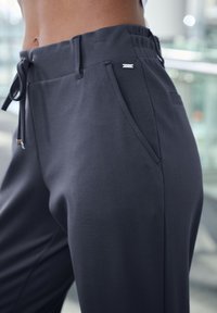 Dunkelgraue Sporthose aus glattem Stoff, mit elastischem Bund und Kordelzug. Verfügt über eine Seitentasche und dezente Markenkennzeichnung in der Nähe der Taille.