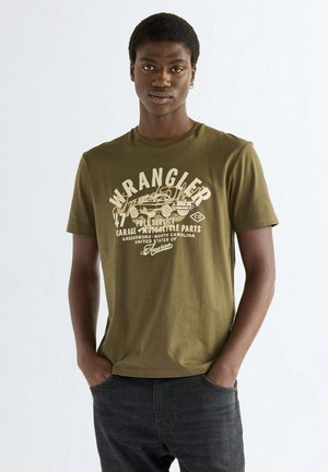 Giovane uomo con capelli corti che indossa una maglietta verde oliva Wrangler con grafica di auto d'epoca e jeans neri, in piedi con le mani nelle tasche.