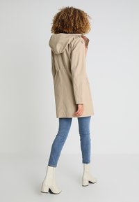Beige lehká parka s kapucí, elastickým pasem, bočními kapsami a hladkým povrchem, kombinovaná s modrými džíny a bílými botami na podpatku.