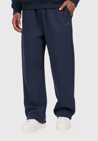 Person in navyblauen Jogginghosen mit "Dropside"-Aufdruck nahe der Tasche, kombiniert mit weißen Sneakers, steht vor einem schlichten Hintergrund.