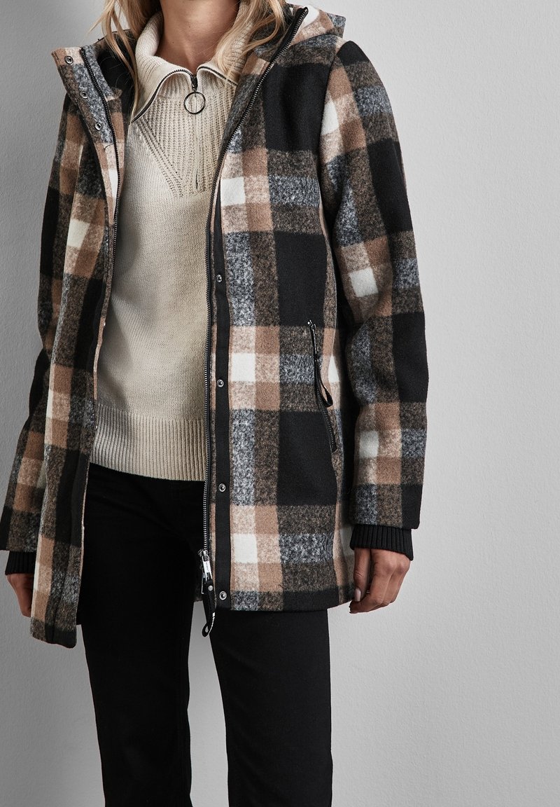 Rutigt svart, brunt och grått ytterplagg med dragkedja, sidofickor och huva. Texturerat tyg och ribbade ärmslut kompletterar designen.