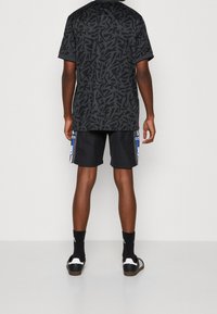 Černé tričko s krátkým rukávem a abstraktním vzorem, spárované s černými šortkami s modrými akcenty a bílým logem Adidas, černé ponožky a sandály.