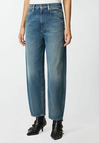 Vaqueros de mezclilla azul oscuro, de corte relajado, de cintura alta con una textura ligeramente desgastada, costuras en contraste y bolsillos frontales visibles, combinados con tacones negros.