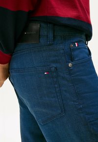 Primer plano de una persona que lleva unos jeans azul oscuro de Tommy Hilfiger y un suéter a rayas rojas y marino, mostrando el bolsillo trasero con el logo de la marca.